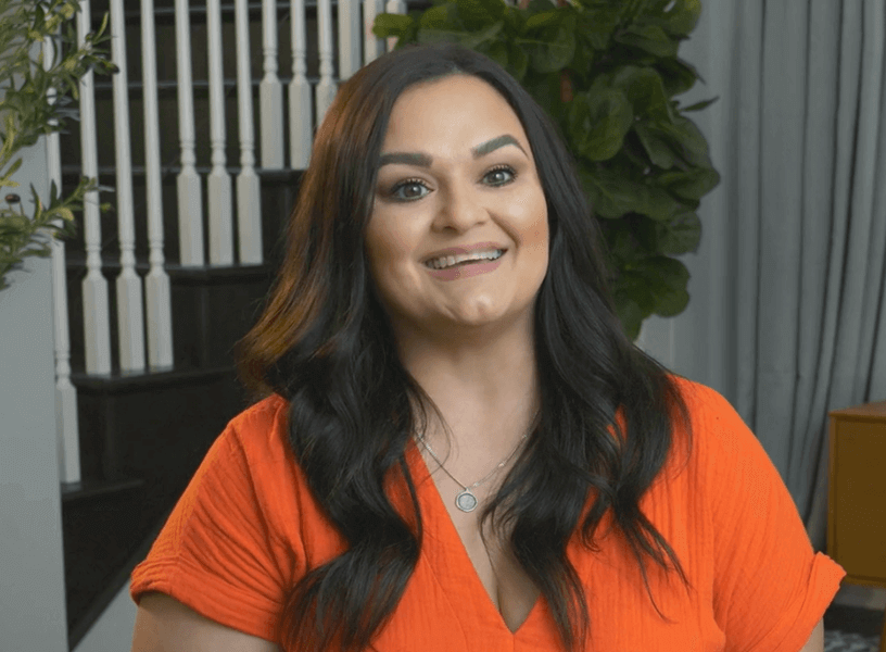 A smiling woman wearing an orange top, with stairs and plants in the background.