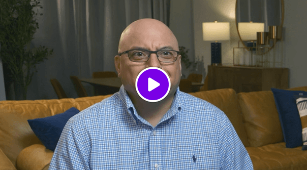Video thumbnail of a man wearing a blue checked shirt and glasses, seated indoors with a lamp, sofa, and plant in the background.