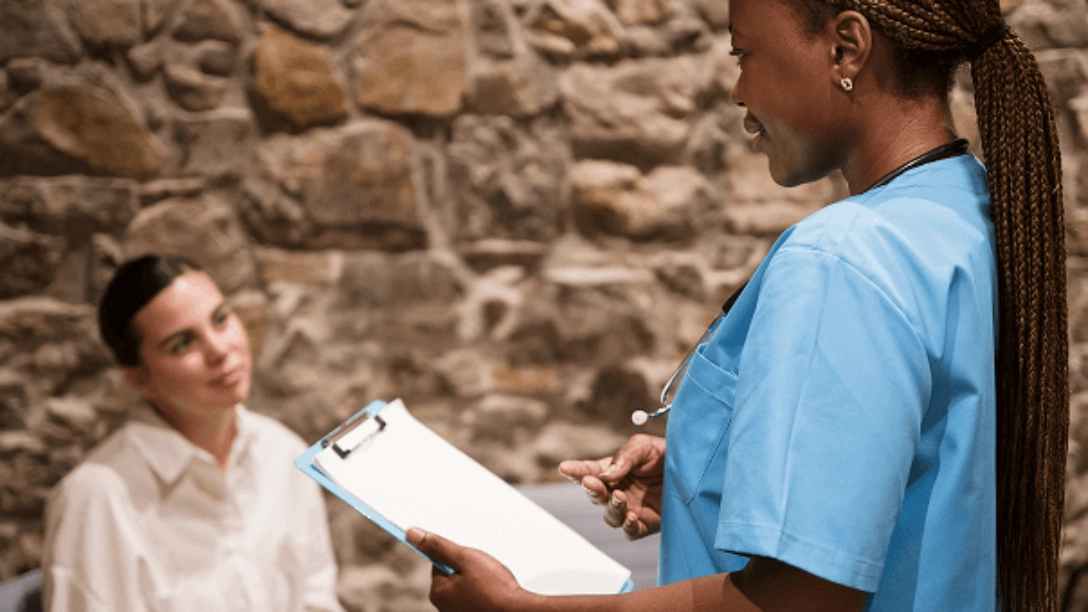 A healthcare professional wearing a blue coat and stethoscope, holding a clipboard talking to a person with a rock wall in the background.