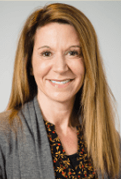 A smiling woman with brown hair, wearing a gray cardigan over a floral-patterned black top, on a gray background.