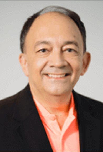 A smiling man with short hair, wearing a black blazer over a pink collared shirt, on a gray background.