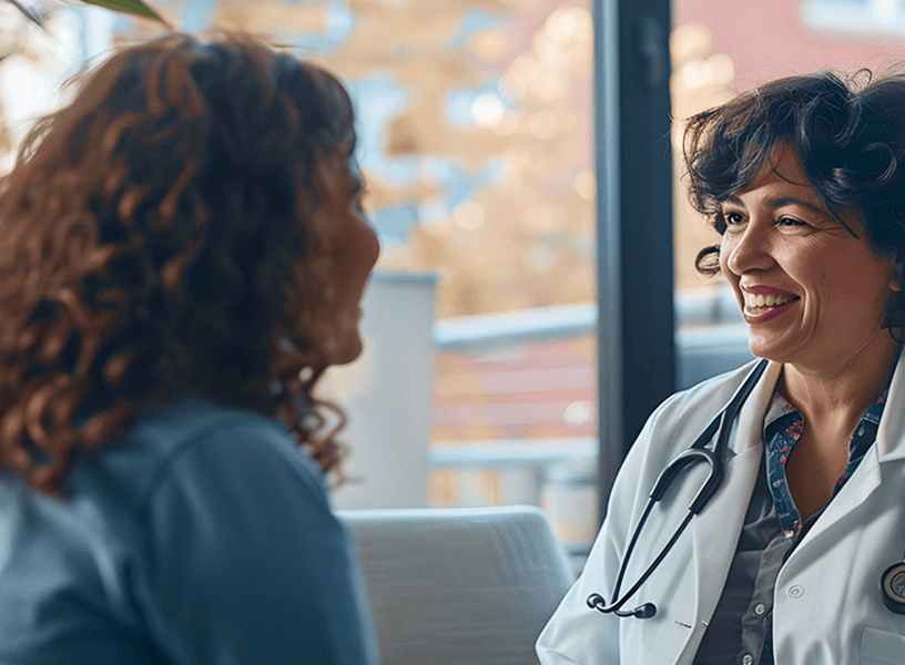 A healthcare professional wearing a white coat and stethoscope talking to a person sitting indoors, with large windows and blurred outdoor scenery in the background.